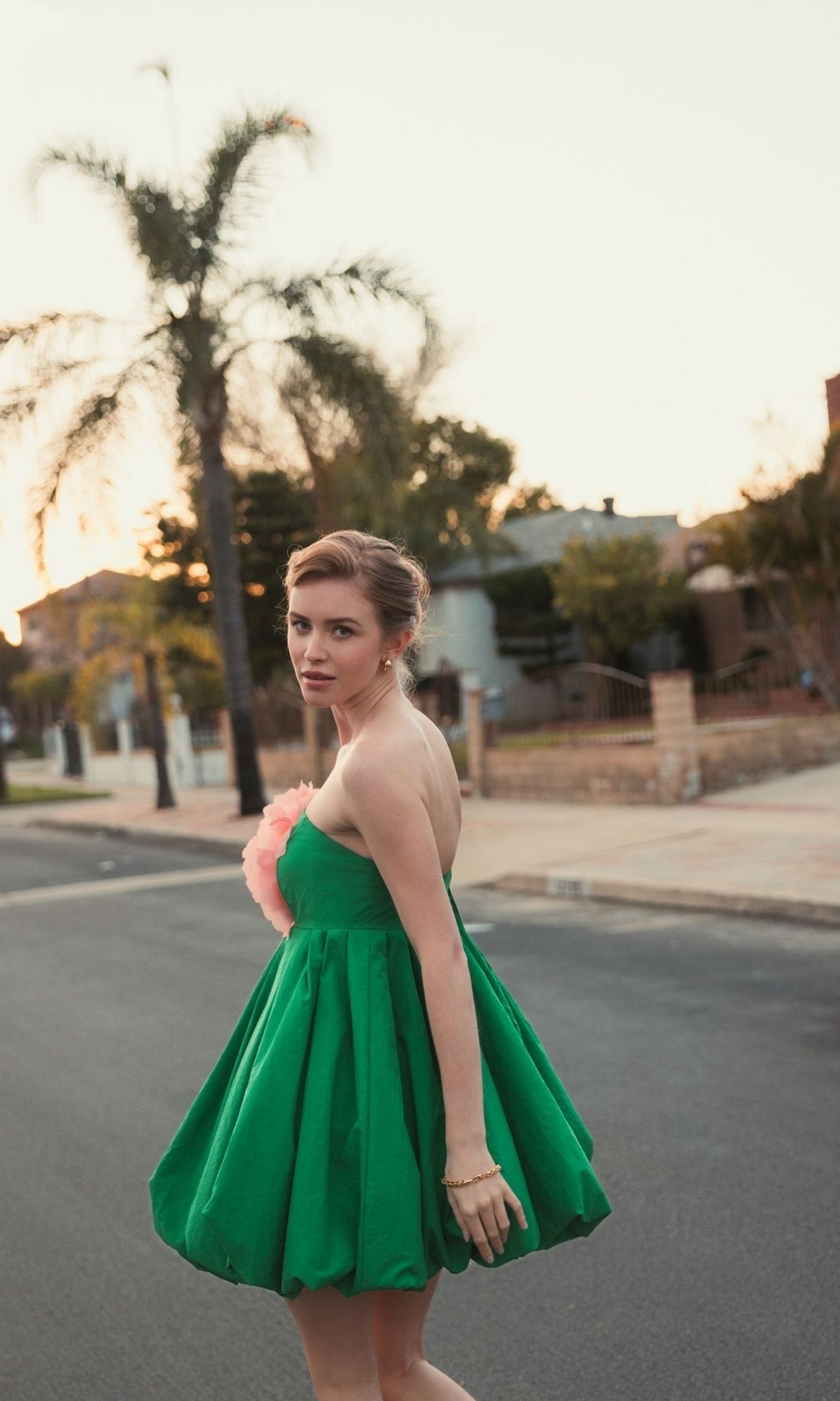 A woman in a Mac Duggal 20931 Short Bubble Party Dress—strapless, green, with a pink flower accent—stands on a residential street at sunset, glancing back over her shoulder.
